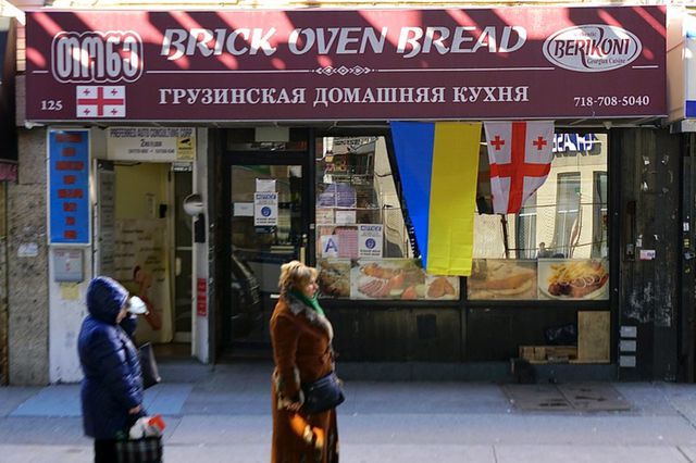 A Ukranian flag and a Georgian flag hang in the front of a bakery in Brighton Beach while two people in winter coats stand on the street in front.